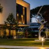 Modern beige building with large glass windows and warm interior lights, set in a landscaped garden at dusk, with white abstract sculptures nearby
