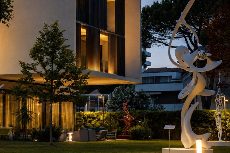 Modern beige building with large glass windows and warm interior lights, set in a landscaped garden at dusk, with white abstract sculptures nearby