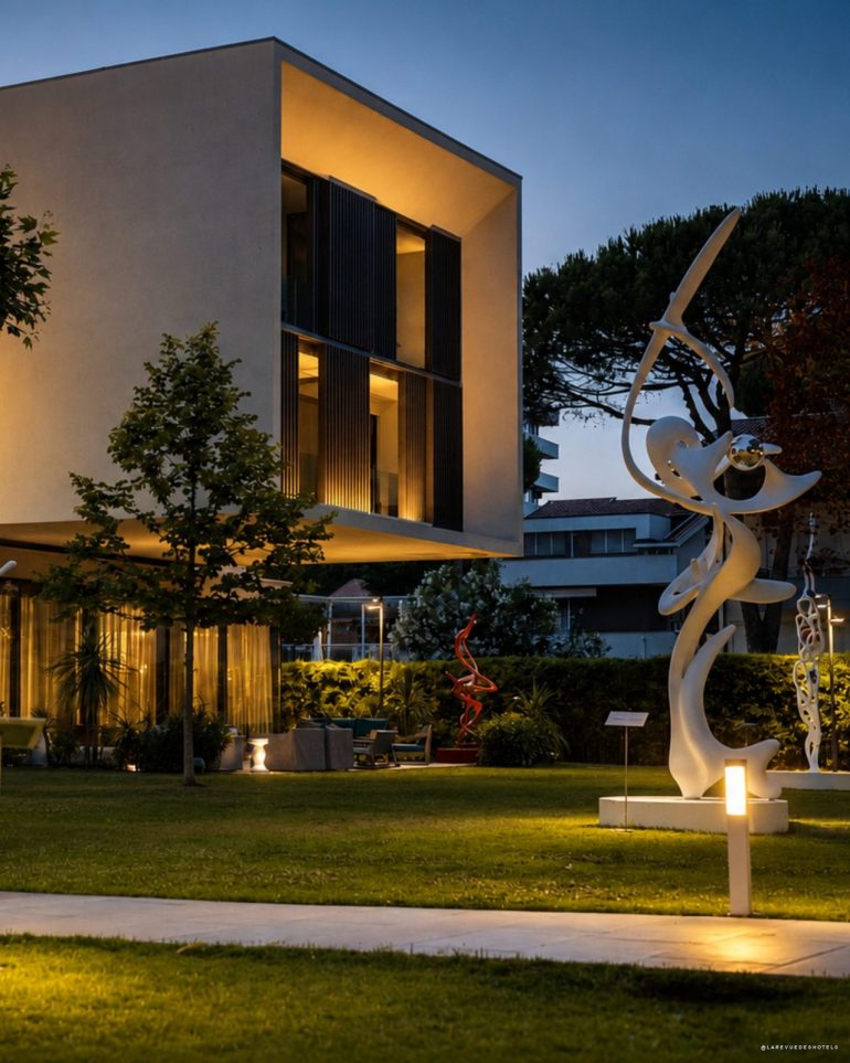 Modern beige building with large glass windows and warm interior lights, set in a landscaped garden at dusk, with white abstract sculptures nearby
