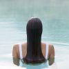 Person with long dark hair in a white swimsuit, viewed from behind, standing chest-deep in a calm swimming pool.