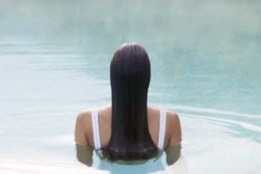 Person with long dark hair in a white swimsuit, viewed from behind, standing chest-deep in a calm swimming pool.