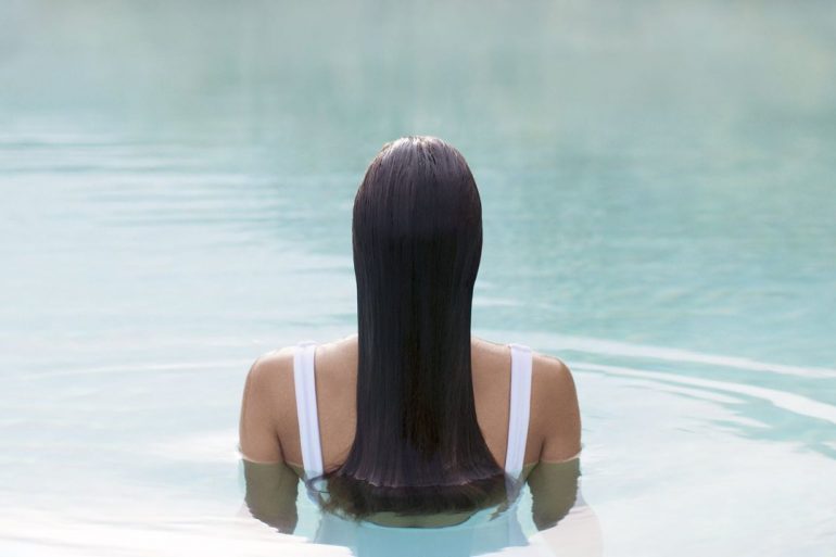 Person with long dark hair in a white swimsuit, viewed from behind, standing chest-deep in a calm swimming pool.