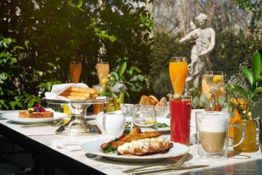 Outdoor brunch table with croissants, baguette sandwiches, pastries, and assorted drinks in a sunlit garden with a statue in the background.