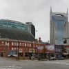 Broad street city block with a red brick historic building on the corner and a tall blue-glass skyscraper behind, under a cloudy sky.