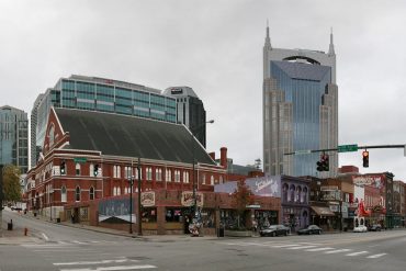 Broad street city block with a red brick historic building on the corner and a tall blue-glass skyscraper behind, under a cloudy sky.