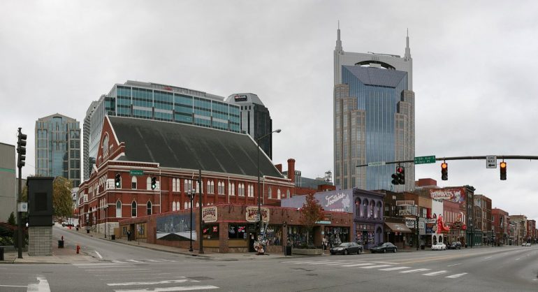 Broad street city block with a red brick historic building on the corner and a tall blue-glass skyscraper behind, under a cloudy sky.