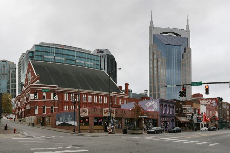 Broad street city block with a red brick historic building on the corner and a tall blue-glass skyscraper behind, under a cloudy sky.