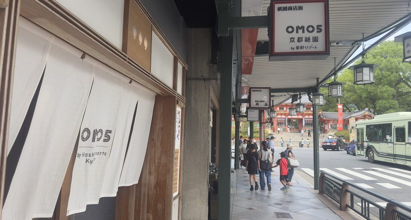 Sidewalk in Kyoto with a white curtain storefront on the left, OM05 sign, pedestrians, and a bus on the road ahead; a traditional temple area visible in the background.