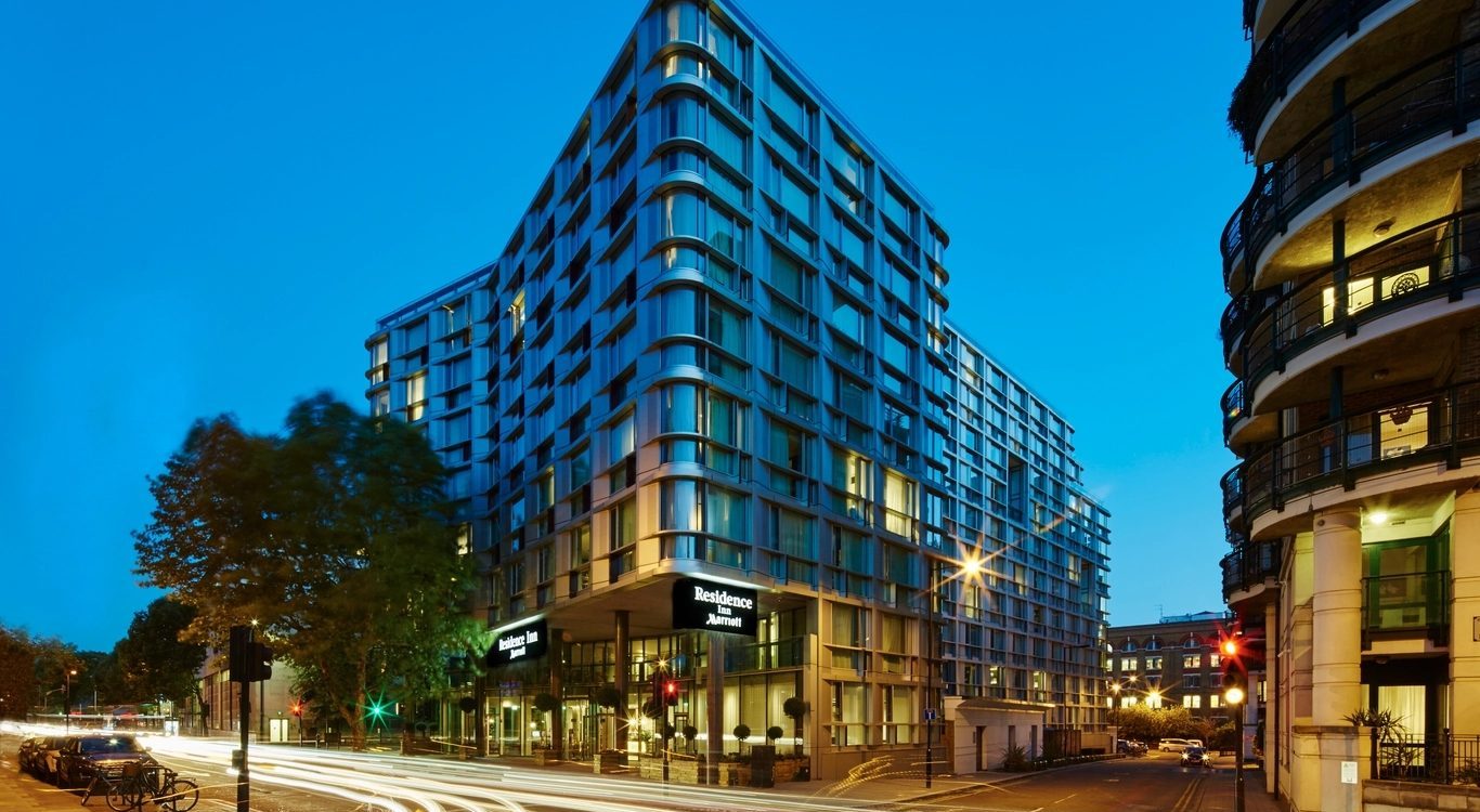 Corner of a modern glass hotel building at dusk with light-trail traffic on the street corner