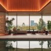 Indoor pool lounge with warm lighting, beige seating, and potted plants, overlooking a city skyline through large glass windows.