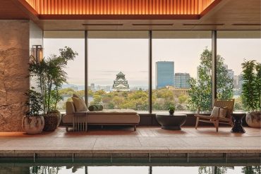 Indoor pool lounge with warm lighting, beige seating, and potted plants, overlooking a city skyline through large glass windows.
