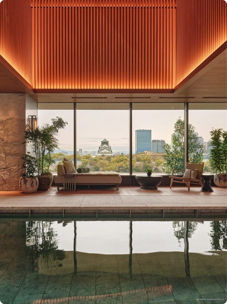 Indoor pool lounge with warm lighting, beige seating, and potted plants, overlooking a city skyline through large glass windows.