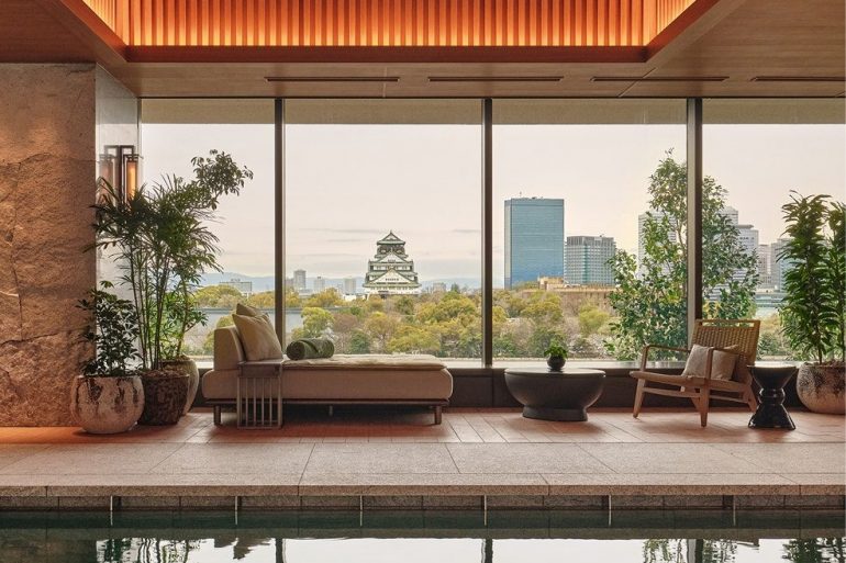 Indoor pool lounge with warm lighting, beige seating, and potted plants, overlooking a city skyline through large glass windows.