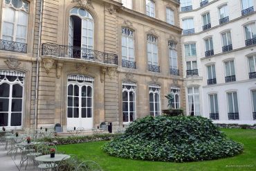 Historic hotel courtyard with a circular ivy-covered fountain statue and wrought-iron tables and chairs on a green lawn.