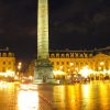 Night view of Place Vendôme in Paris with a tall obelisk at center, surrounding neoclassical buildings, and glowing street lamps reflecting on wet cobblestones.