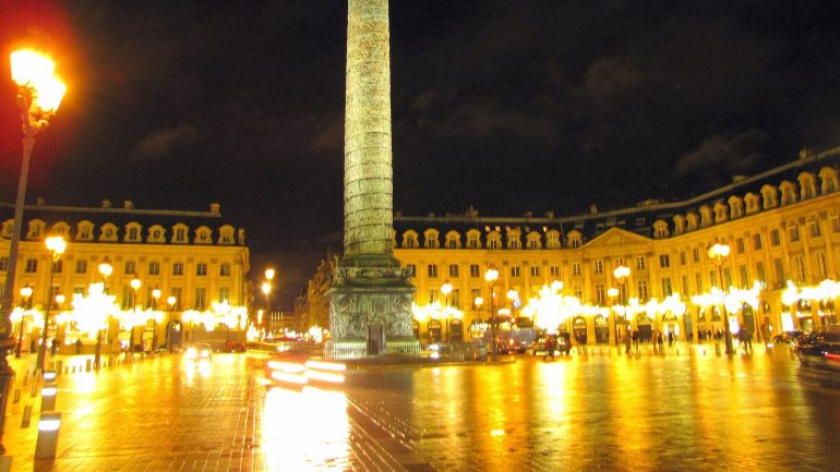 Night view of Place Vendôme in Paris with a tall obelisk at center, surrounding neoclassical buildings, and glowing street lamps reflecting on wet cobblestones.