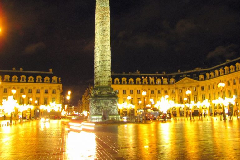 Night view of Place Vendôme in Paris with a tall obelisk at center, surrounding neoclassical buildings, and glowing street lamps reflecting on wet cobblestones.