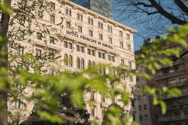Facade of the Hotel Principe di Savoia with arched windows, framed by green tree branches and a blue sky above.