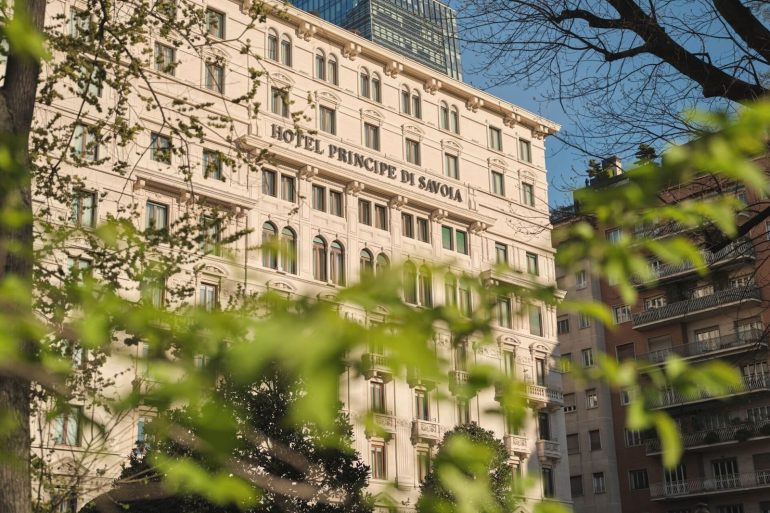 Facade of the Hotel Principe di Savoia with arched windows, framed by green tree branches and a blue sky above.