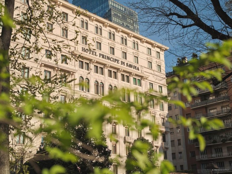 Facade of the Hotel Principe di Savoia with arched windows, framed by green tree branches and a blue sky above.
