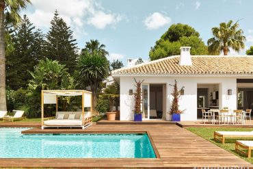 Poolside wooden deck with a white canopy daybed, lounge chairs, and a tropical garden next to a white villa with a tiled roof.