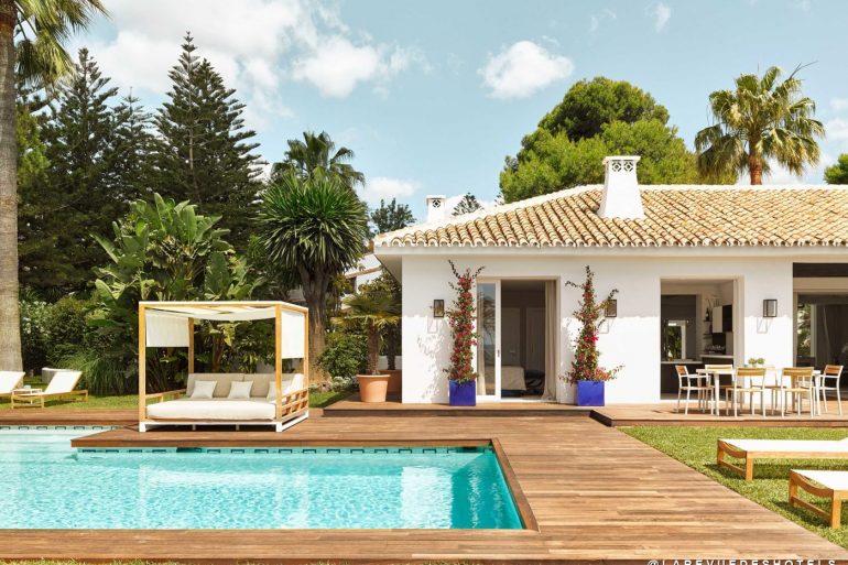 Poolside wooden deck with a white canopy daybed, lounge chairs, and a tropical garden next to a white villa with a tiled roof.
