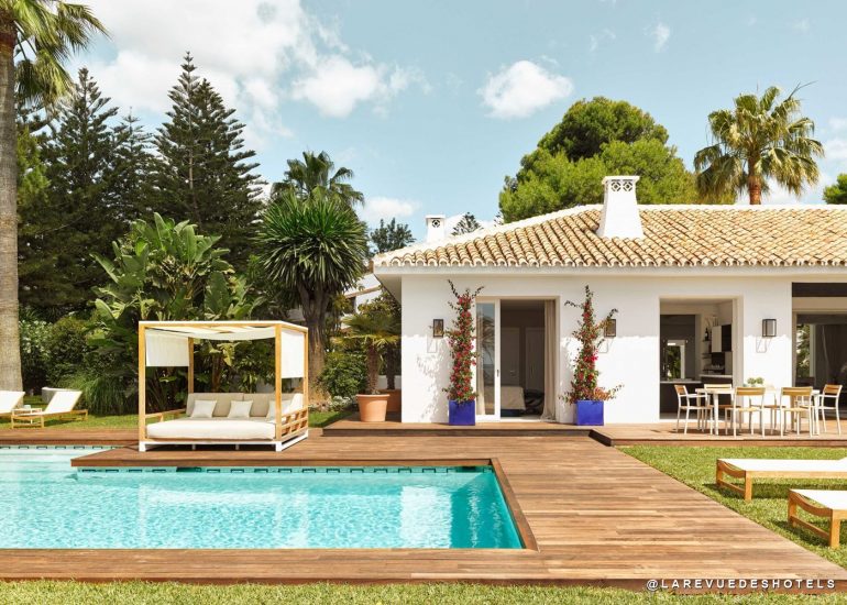 Poolside wooden deck with a white canopy daybed, lounge chairs, and a tropical garden next to a white villa with a tiled roof.
