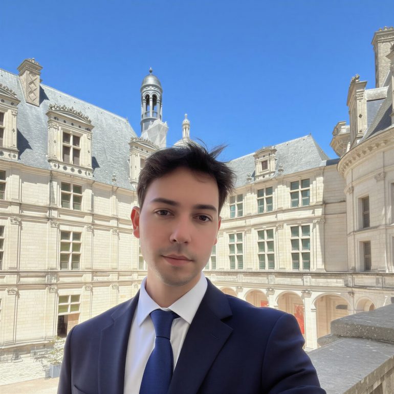 Man in a navy suit and blue tie taking a selfie in a historic European courtyard with ornate stone buildings and a clear blue sky.