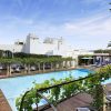 Azure pool surrounded by wooden decking and white Mediterranean buildings, with lounge chairs and greenery.