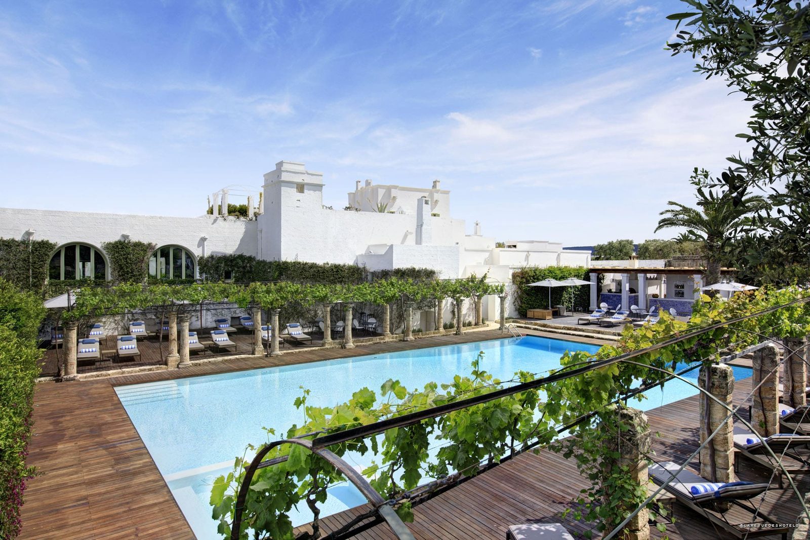 Azure pool surrounded by wooden decking and white Mediterranean buildings, with lounge chairs and greenery.