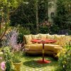 Cosy yellow sectional sofa around a red round table in a lush garden with blooming plants and greenery.