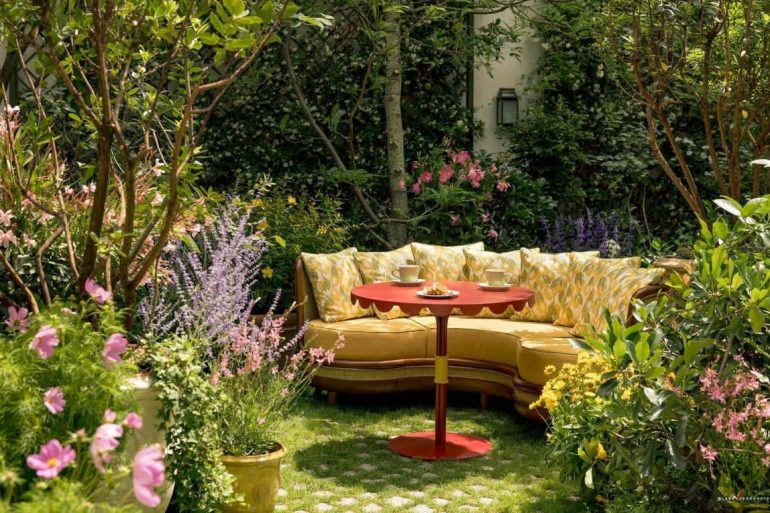 Cosy yellow sectional sofa around a red round table in a lush garden with blooming plants and greenery.
