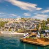 Resort along a turquoise bay with white hillside buildings, palm trees, and wooden decks with lounge chairs.