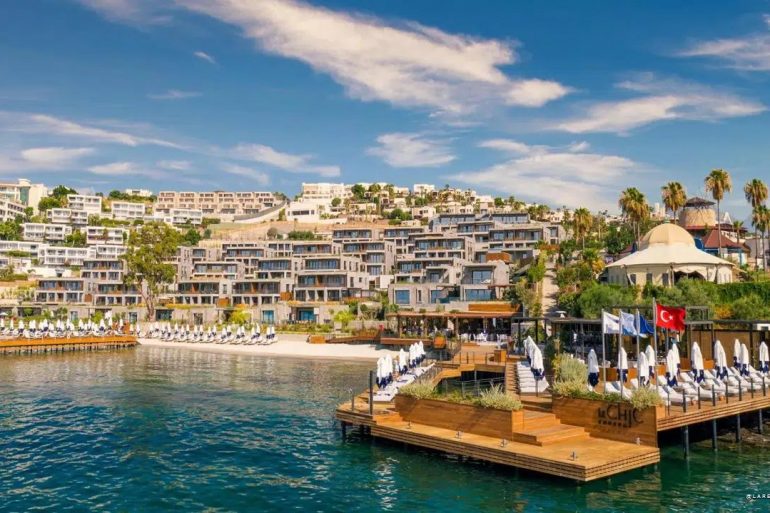 Resort along a turquoise bay with white hillside buildings, palm trees, and wooden decks with lounge chairs.