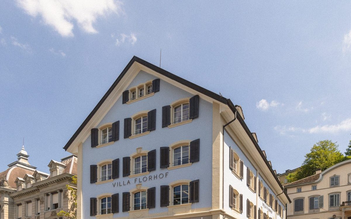 Blue and beige hotel building with black shutters, labeled Villa Florhof, on a cobblestone street against a clear blue sky.
