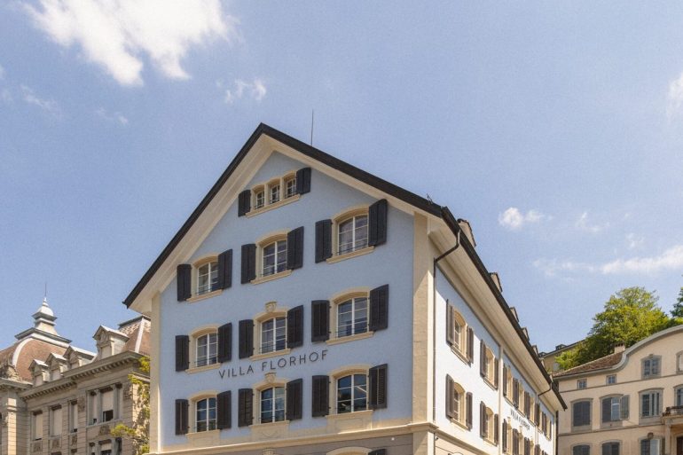 Blue and beige hotel building with black shutters, labeled Villa Florhof, on a cobblestone street against a clear blue sky.
