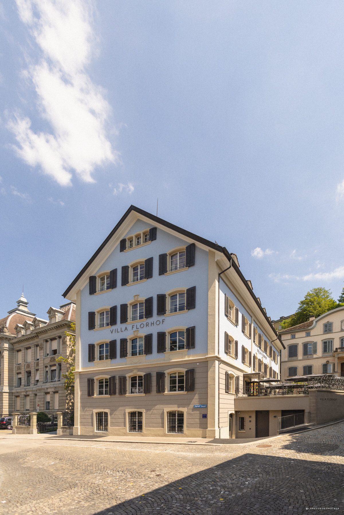 Blue and beige hotel building with black shutters, labeled Villa Florhof, on a cobblestone street against a clear blue sky.