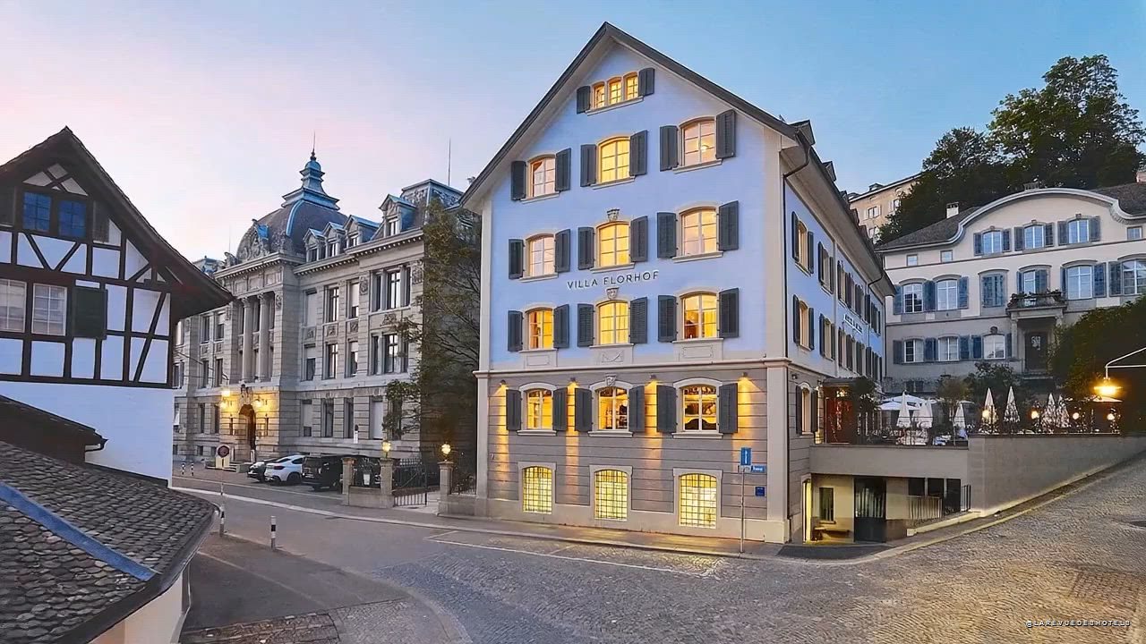 Front view of a European hotel (Villa Florhof) on a cobblestone street, warm lights glow from windows at dusk.