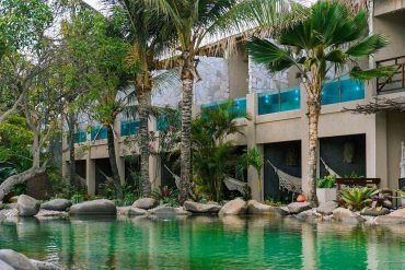 Resort courtyard with a turquoise pool, palm trees, hammocks, and a stone-clad building facade.