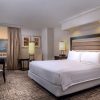 Hotel room with a king bed, padded headboard, two nightstands with lamps, a desk and chair, and a seating area by a window overlooking the city.