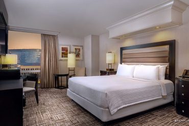 Hotel room with a king bed, padded headboard, two nightstands with lamps, a desk and chair, and a seating area by a window overlooking the city.
