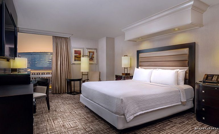 Hotel room with a king bed, padded headboard, two nightstands with lamps, a desk and chair, and a seating area by a window overlooking the city.