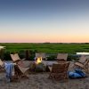 Wicker chairs arranged in a circle around a campfire on a sandy beach, marsh grass and a sunset horizon in the distance.