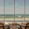 Indoor seating area facing a glass wall with a scenic beach and ocean beyond, sunlight streaming on cushions and a round table set for two