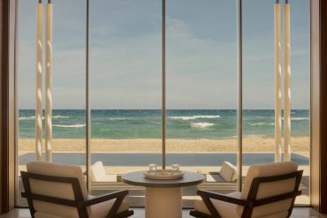 Indoor seating area facing a glass wall with a scenic beach and ocean beyond, sunlight streaming on cushions and a round table set for two
