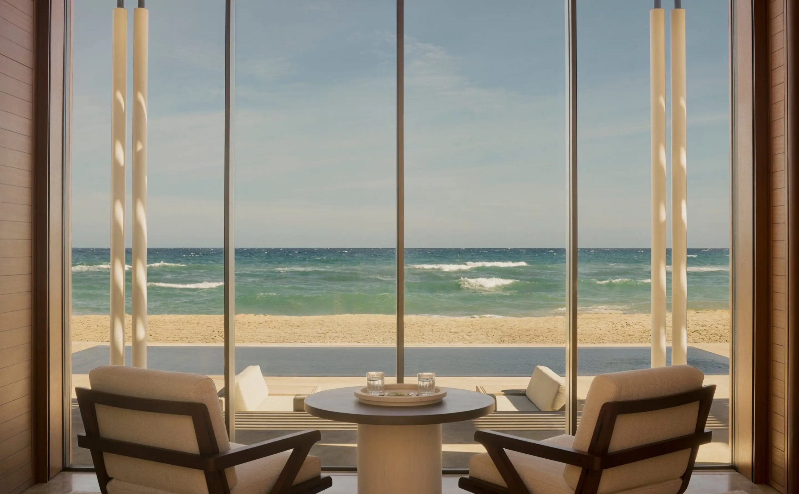 Indoor seating area facing a glass wall with a scenic beach and ocean beyond, sunlight streaming on cushions and a round table set for two