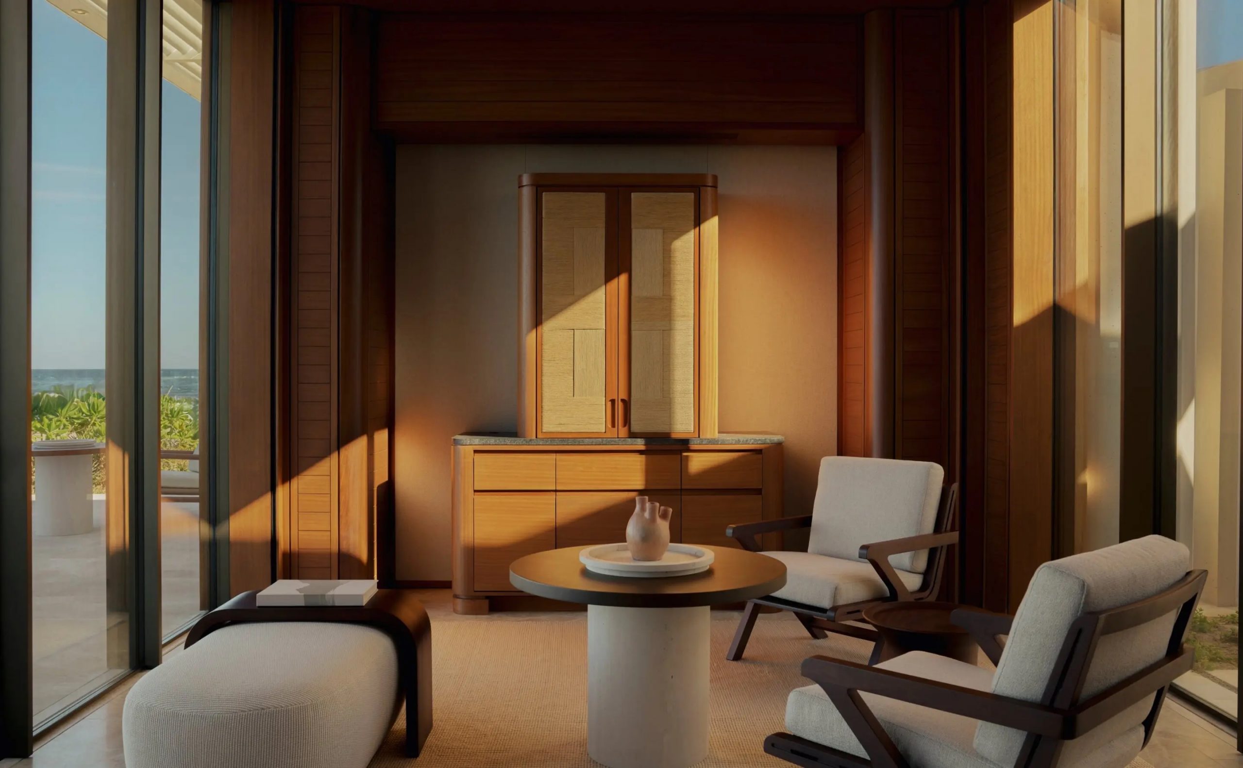 Indoor seating area facing a glass wall with a scenic beach and ocean beyond, sunlight streaming on cushions and a round table set for two