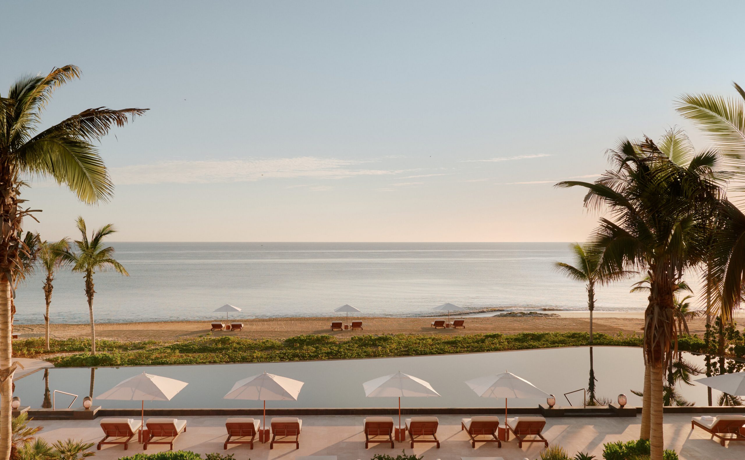 Indoor seating area facing a glass wall with a scenic beach and ocean beyond, sunlight streaming on cushions and a round table set for two