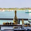 Champagne bottle centerpiece on a table with desserts and wine glasses, overlooking turquoise sea and boats in the distance.
