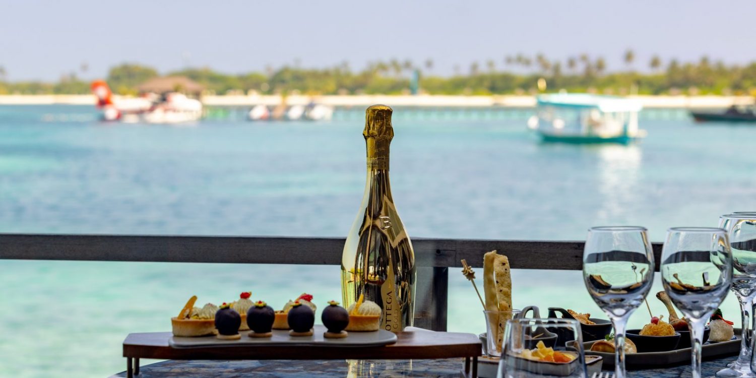 Champagne bottle centerpiece on a table with desserts and wine glasses, overlooking turquoise sea and boats in the distance.